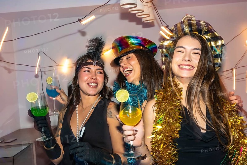 Three young adult women friends enjoying a lively new year's eve or costume party, laughing together while holding colorful cocktails at a festive home celebration