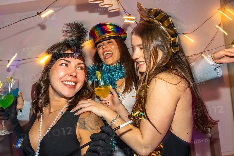 Young women in festive costumes laugh and toast cocktails at a colorful new year's eve home party, sharing joyful friendship, confetti, and lively celebration together