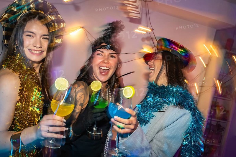Diverse women in festive party hats and tinsel clink colorful lime garnished cocktails, laughing and dancing at a vibrant, energetic new years eve or costume party celebration