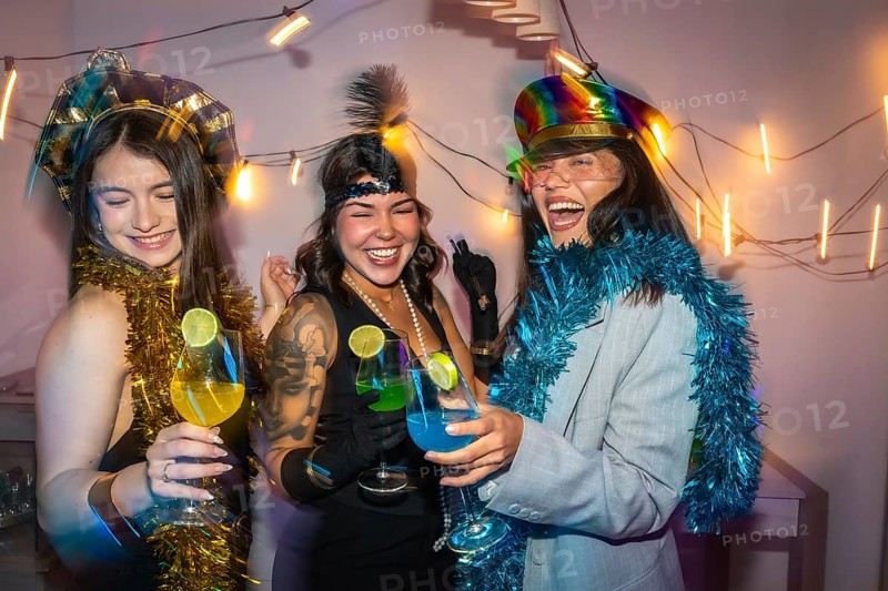 Three young women friends enjoying a fun new year's eve party, celebrating with colorful cocktails, dressing up in costume hats and festive tinsel while laughing together
