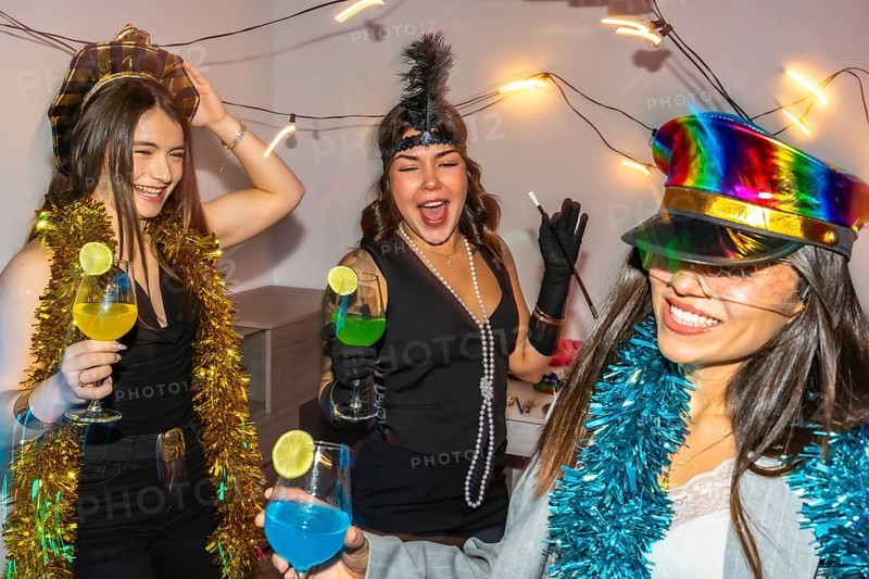 Young women friends celebrating new year's eve or a costume party indoors, laughing and dancing while holding colorful cocktails, surrounded by festive decorations and lights