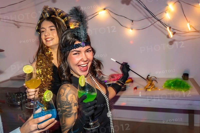 Young women in 1920s style costumes celebrating at home with cocktails, string lights and disco ball sparkle, laughing and enjoying a festive, vintage themed party with friends