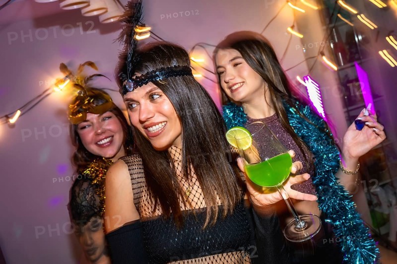 Three friends smiling and celebrating a festive costume party at home, holding a green cocktail and enjoying the vibrant atmosphere with decorative lights