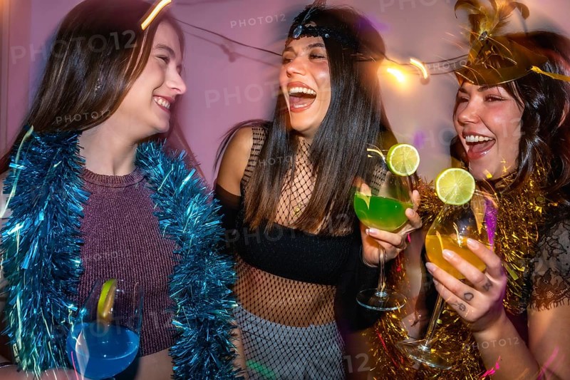 Group of women laughing and holding cocktails while celebrating with party decorations, enjoying a fun night out or costume party with friends, full of joy and friendship