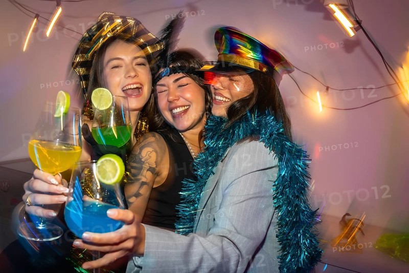 Three happy young women in festive costumes laughing and cheering with vibrant cocktails, enjoying a fun celebration together in a lively party atmosphere