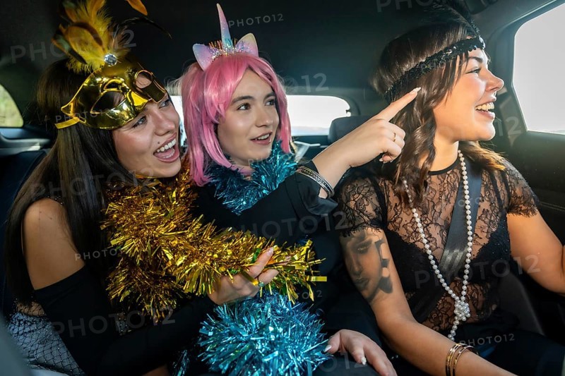 Three young women dressed in cheerful costumes with tinsel on the back seats of a car, joyfully interacting and laughing while pointing at something outside