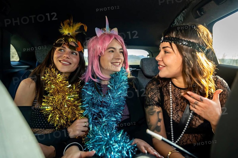 Three young women in colorful costume accessories and party attire laughing and talking in the back seat of a car, celebrating a special event together