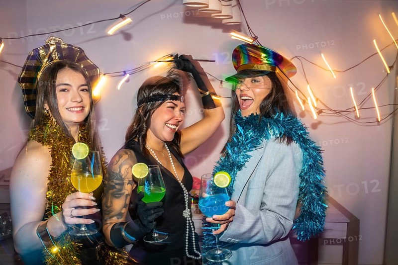 Three diverse women wearing festive costumes, decorations, and holding colorful cocktails, enjoying a lively friends costume party at home, celebrating an event