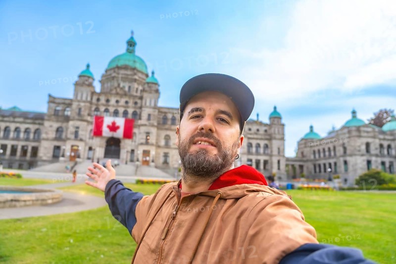 Happy bearded tourist taking a selfie with outstretched arm in front of the parliament buildings in victoria, british columbia, with canadian flag displayed prominently