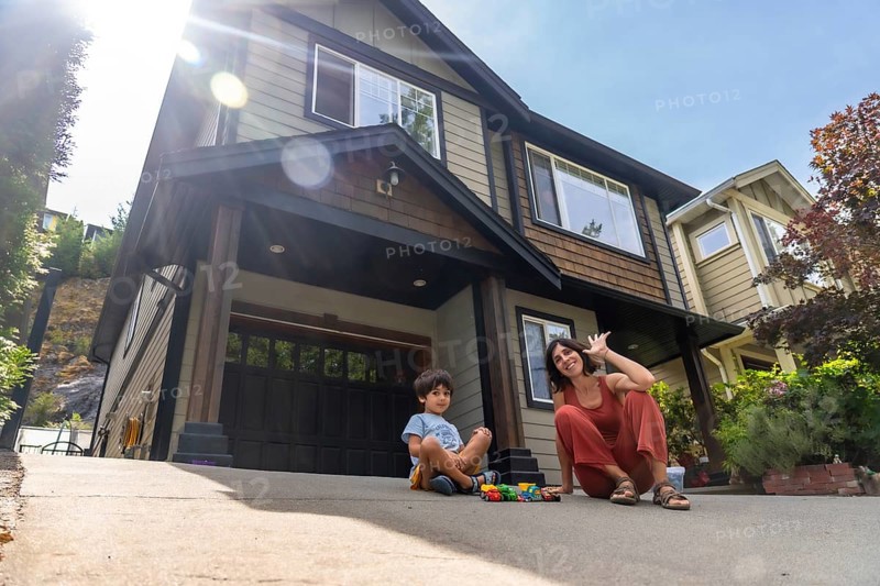 Mother and son are sitting on their driveway in front of their house, enjoying quality time together on a bright summer day, waving and playing with toy cars