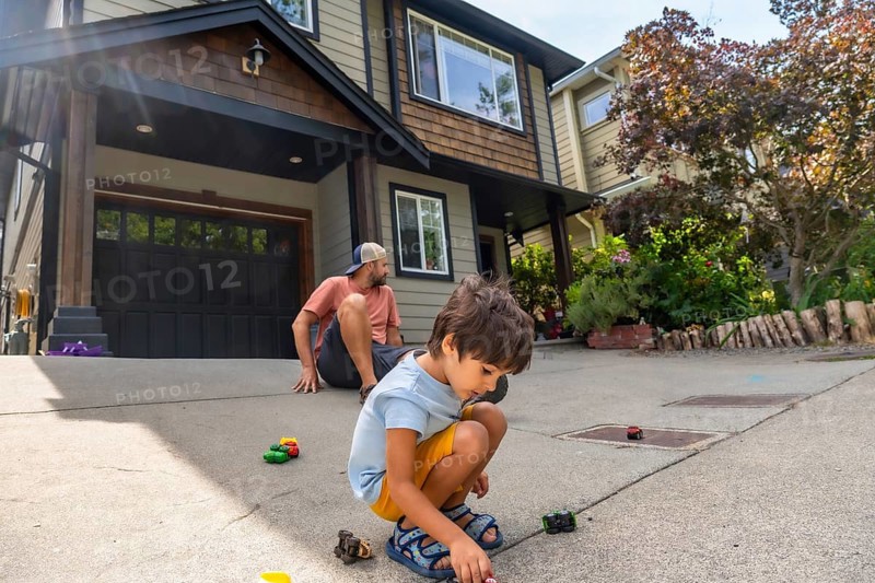 Young boy plays with toy cars on the driveway of a modern home while his father watches from behind, creating a heartwarming scene of family bonding and childhood joy