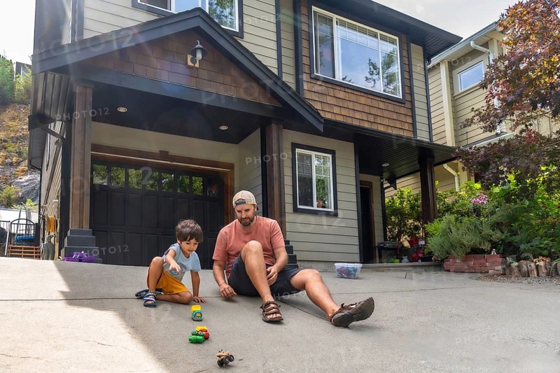 Father and son are sitting on their driveway, playing with a collection of toy cars in front of their suburban home on a sunny summer day