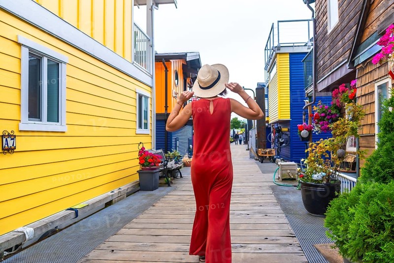 Tourist wearing a straw hat and red dress walking along the wooden boardwalk at fisherman's wharf park in victoria, british columbia, enjoying the colorful floating houses and vibrant flowers