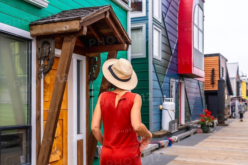 Female tourist wearing a straw hat and a red dress is enjoying a summer day walking down the fisherman's wharf park in victoria, british columbia, canada, admiring the colorful floating houses