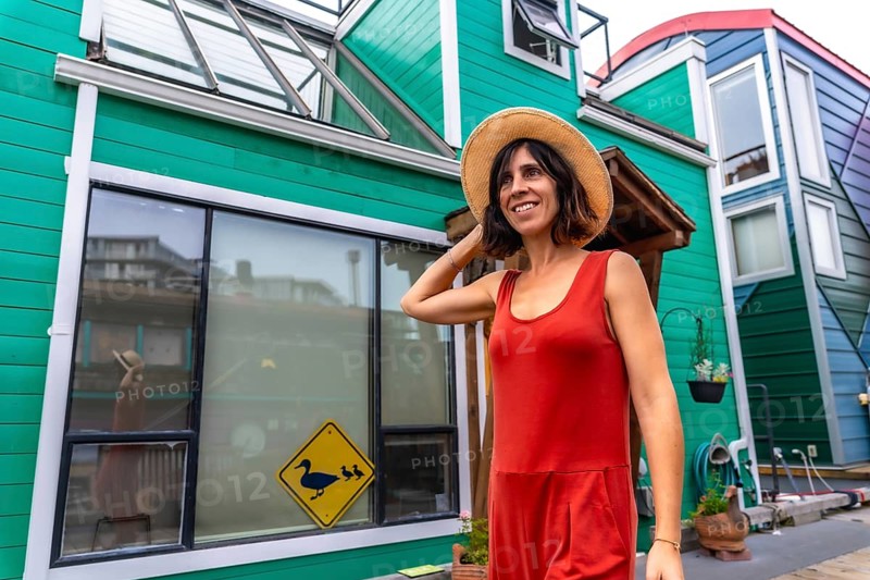 Happy tourist adjusting her straw hat while walking and smiling in front of colorful floating houses in fisherman's wharf park in victoria, british columbia