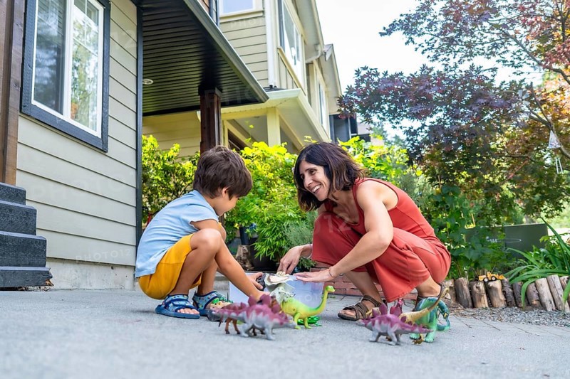 Mother and her son are happily playing with a collection of dinosaur toys on the driveway in front of their house, enjoying quality time together