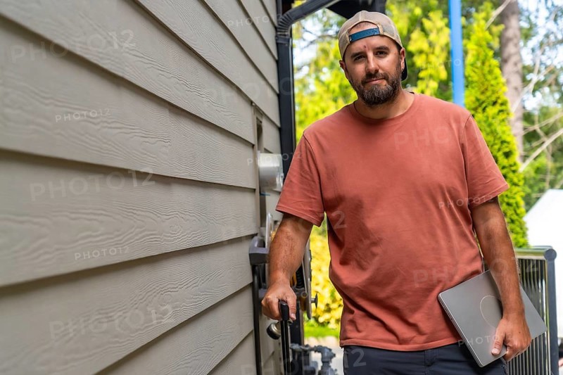 Maintenance worker walking alongside a residential building, carrying a laptop and various tools, dressed in casual attire with a cap, ready for service and repair tasks