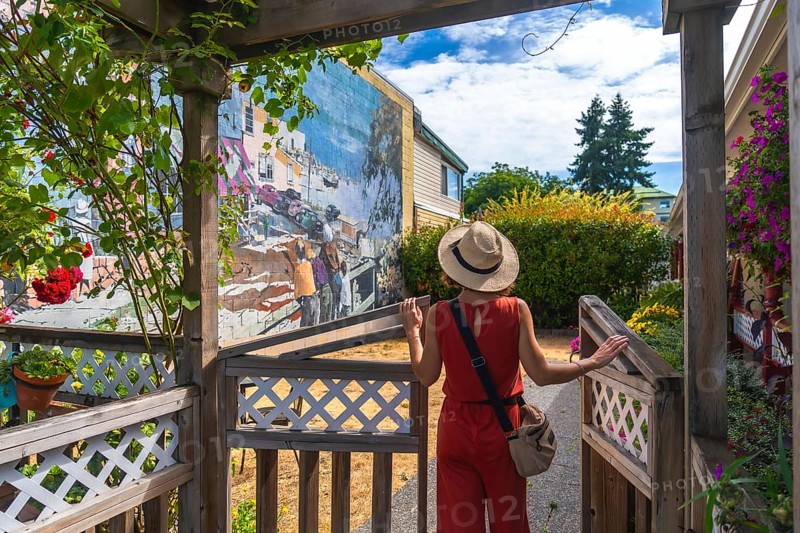Female tourist in a straw hat and red jumpsuit opening a wooden gate to admire a vibrant mural depicting the city's history in chemainus, vancouver island, on a sunny summer day