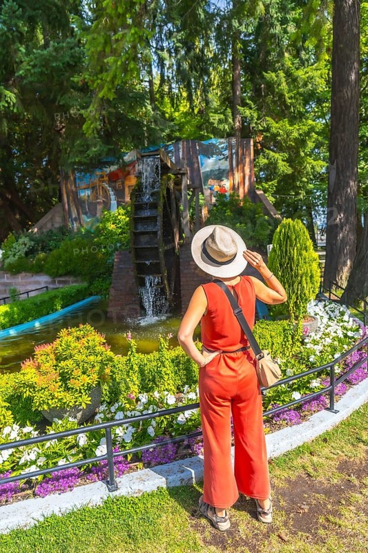 Female tourist wearing a straw hat and a jumpsuit is admiring a watermill and a colorful mural in a park in chemainus, british columbia, during summer