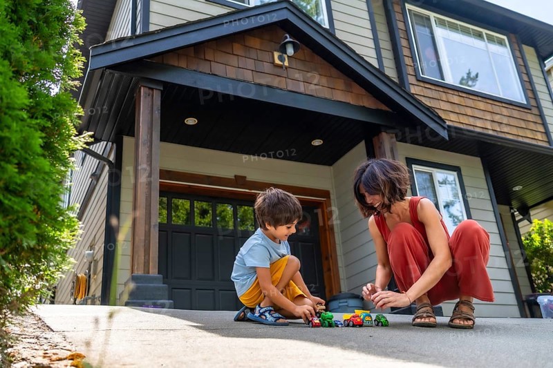Mother and her young son are happily playing with toy cars on the driveway of their suburban home, enjoying quality time together on a sunny day