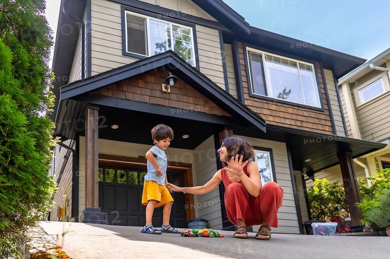Mother interacts with her young son, playing with toy cars on the driveway of their modern suburban home, creating a heartwarming scene of family life and childhood joy