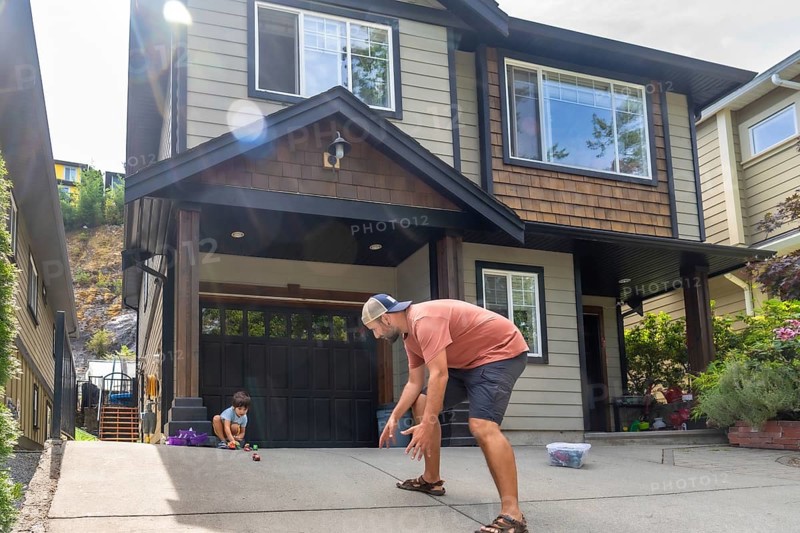 Father crouching down and playing with toy cars alongside his young son in the driveway of their modern two story home, creating joyful moments of bonding and fun