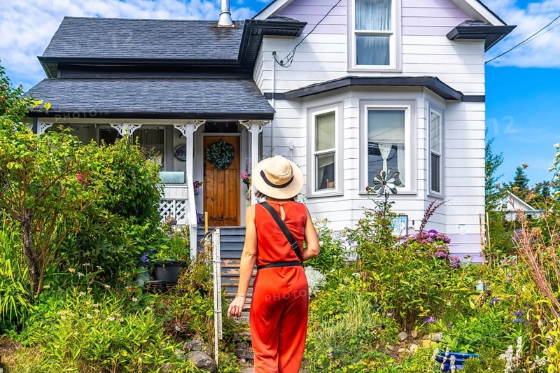 Tourist wearing a straw hat and an orange jumpsuit walks up a garden path towards a charming house in chemainus, british columbia, canada, on a sunny summer day