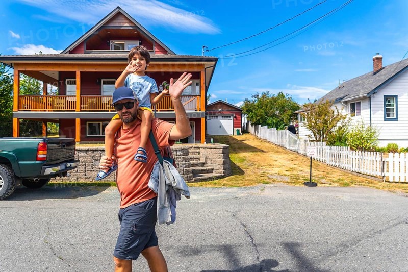 Father carrying his son on his shoulders waving in front of their house in chemainus, vancouver island, british columbia, canada, during a sunny summer day