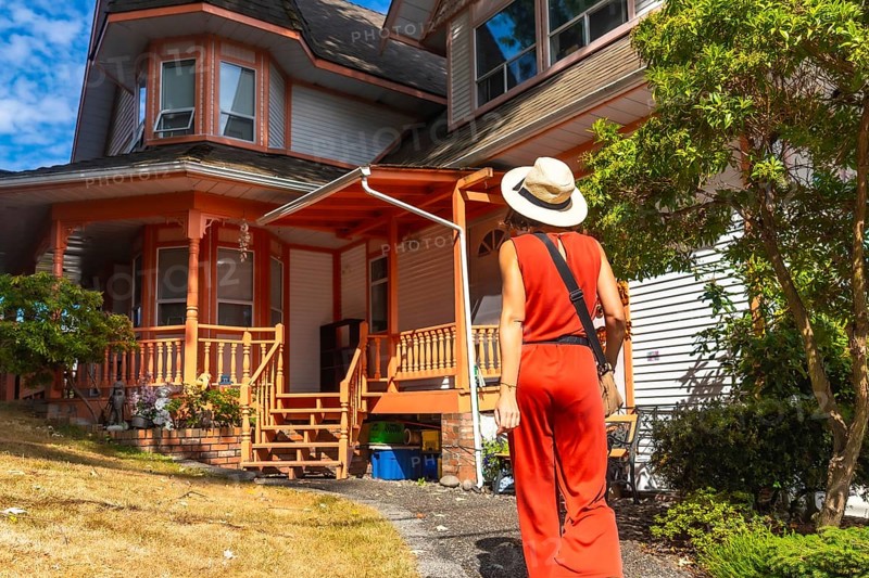 Tourist wearing a straw hat and a red jumpsuit is walking on a path near a traditional wooden house with an orange porch in chemainus, vancouver island, during a sunny summer day