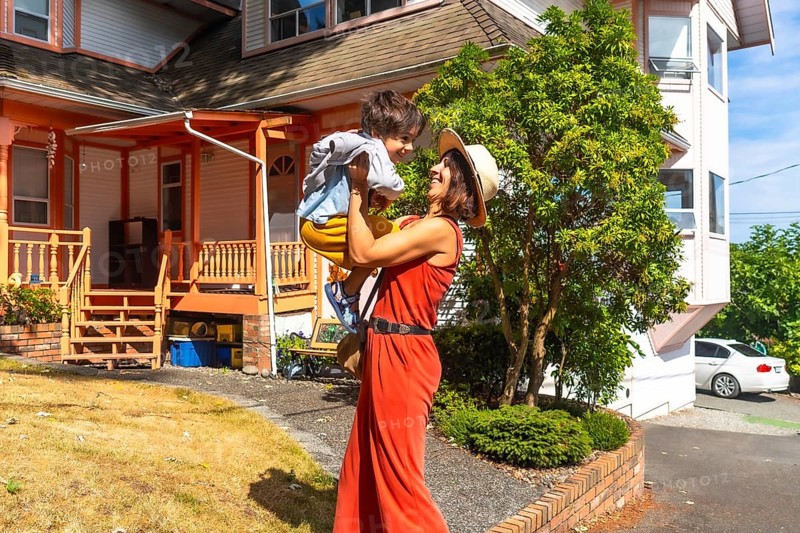 Happy mother wearing a straw hat and an orange jumpsuit lifting up her cheerful son in the yard of their house in chemainus, british columbia, canada