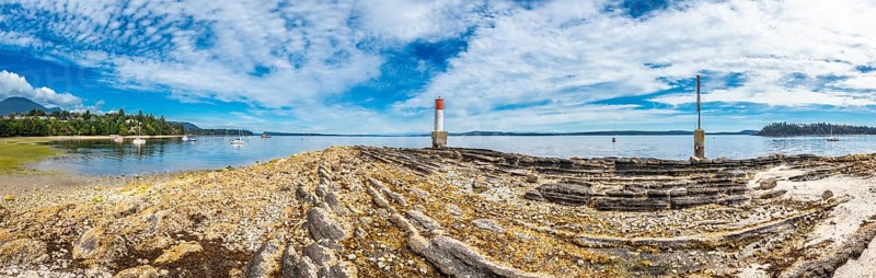 Rocky coastline stretches towards a red and white lighthouse, with sailboats dotting the tranquil waters of chemainus bay under a bright summer sky on vancouver island, british columbia