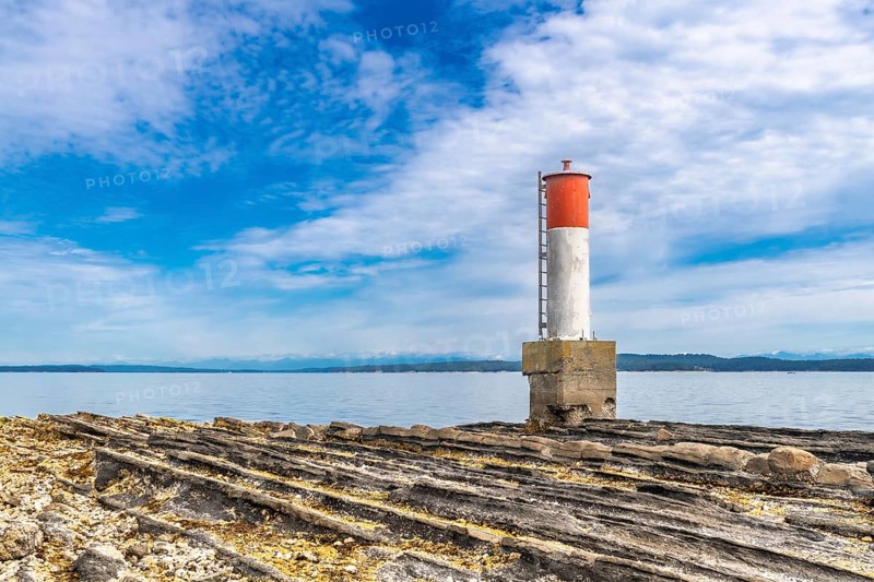 Red and white navigation marker stands on a rocky coastline in chemainus, british columbia, with calm waters and distant mountains under a blue sky with white clouds