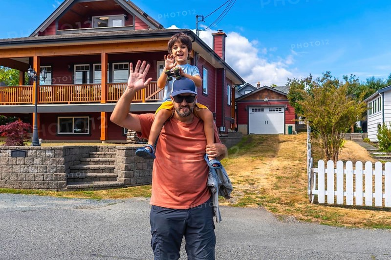 Father carrying his son on his shoulders, both waving, standing in front of their house in chemainus, vancouver island, british columbia, canada, enjoying a summer day
