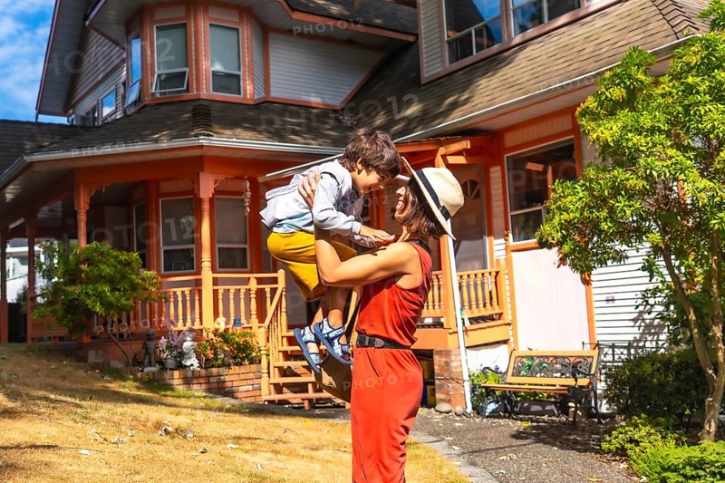 Happy mother lifting her son in the air, having fun in the yard of their colorful house in chemainus, vancouver island, british columbia, canada, during a sunny summer day