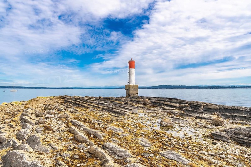 Red and white lighthouse rising from a rocky, textured coastline in chemainus, vancouver island, british columbia, canada, creating a picturesque maritime scene under a cloudy summer sky