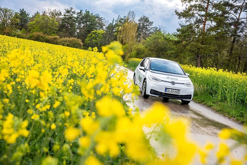 White car drives on a narrow road through a blooming yellow field in rural surroundings, ID3 VW, deer car sharing. Calw, Germany