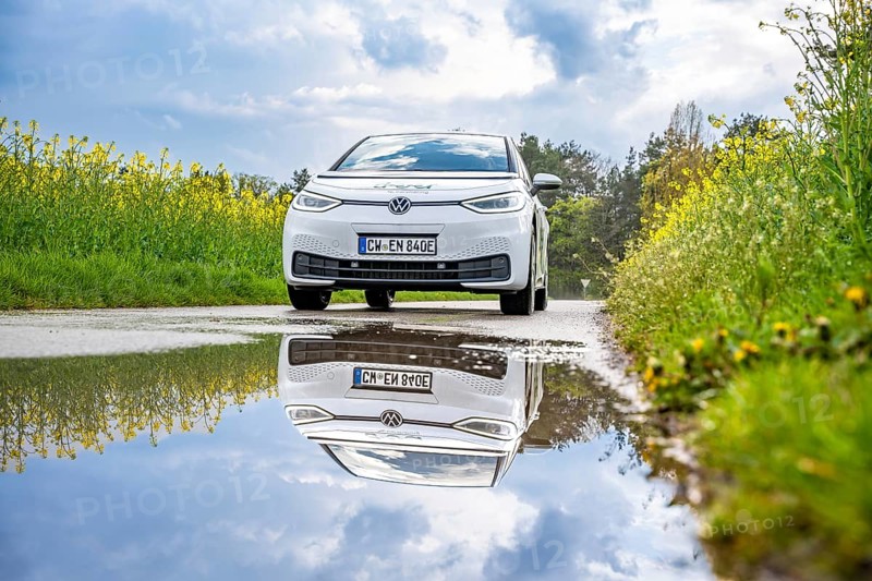 A white electric car on a country road, reflected in a puddle, surrounded by yellow flowers and blooming nature, ID3 VW, deer car sharing. Calw, Germany