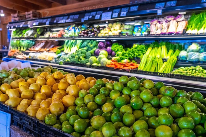 Fresh limes and lemons are piled high in the foreground, with a colorful display of various fruits and vegetables arranged on shelves in the background of a grocery store produce section