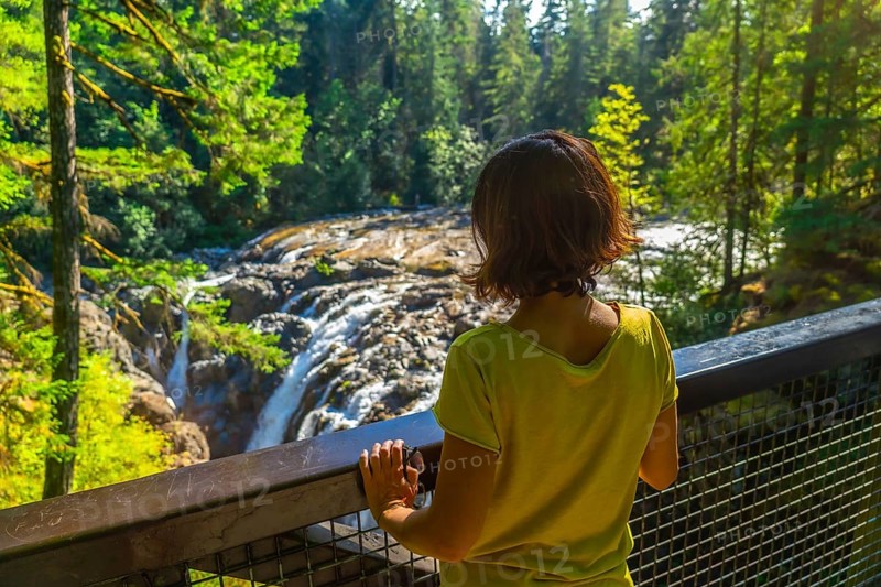 Female tourist standing on a viewpoint, admiring englishman river falls cascading through lush green forest in vancouver island, british columbia, canada, during a sunny summer day
