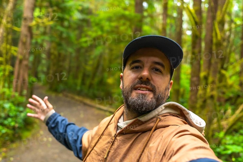 Tourist wearing casual clothing and a baseball cap is gesturing to the rainforest trail while taking a selfie in ucluelet on vancouver island, british columbia, canada