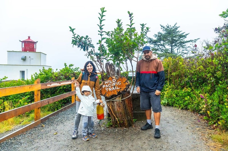 Happy family posing near the wild pacific trail sign with amphitrite point lighthouse in the background, enjoying a cloudy summer day in ucluelet, british columbia, canada