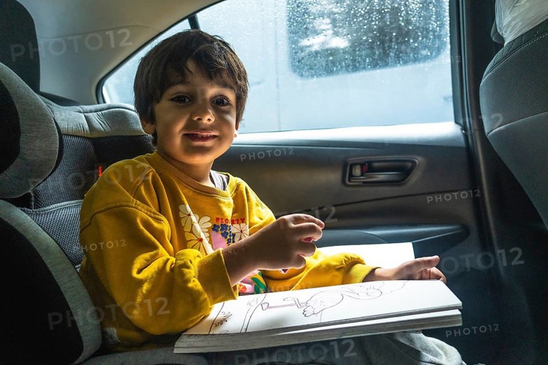 Young child happily drawing in a coloring book while secured in a car seat during a road trip, providing entertainment and creative expression on the journey