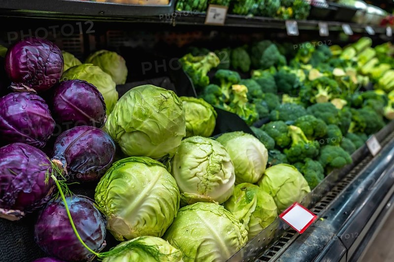 Fresh green and red cabbages and broccoli heads are neatly arranged on a refrigerated shelf in a supermarket, offering a colorful and healthy selection for shoppers seeking fresh produce