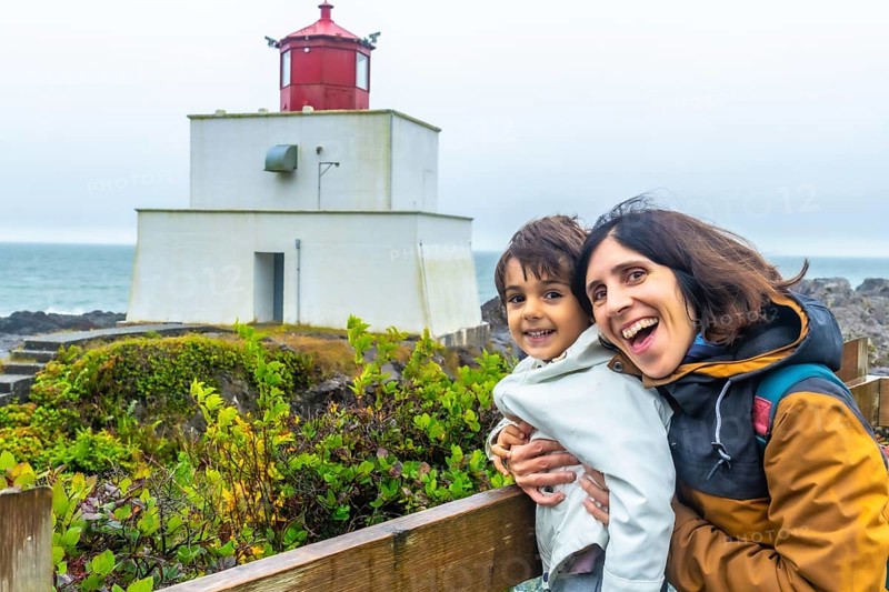 Happy mother and child hugging and smiling with the amphitrite point lighthouse in ucluelet, british columbia, canada, in the background, enjoying a cloudy day by the pacific ocean