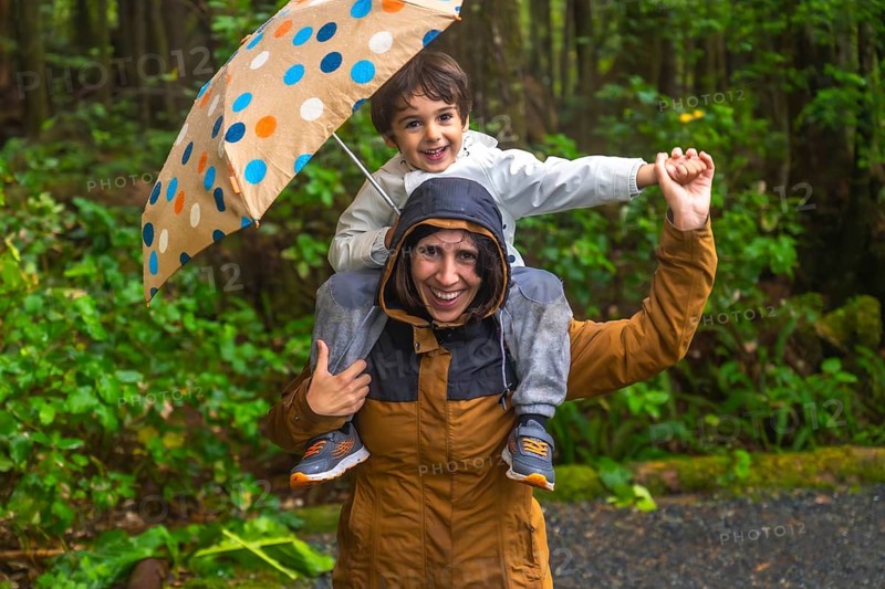 Mother carries her smiling son on her shoulders under a colorful umbrella, both wearing raincoats and enjoying a rainy day adventure in a lush green forest