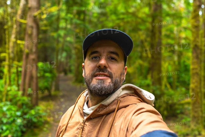 Tourist in a cap and light brown jacket taking a selfie while walking along a lush rainforest trail in ucluelet, british columbia, surrounded by natural beauty