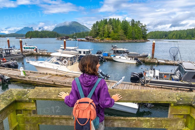 Female tourist with backpack contemplating moored boats in tofino harbor, vancouver island, british columbia, canada, during a sunny summer day with clouds and mountains in the background