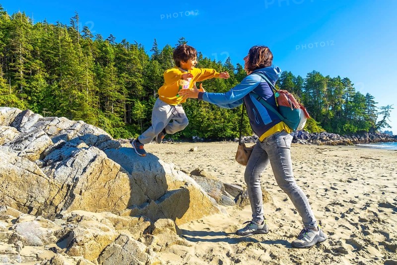 Joyful child leaping from a rock into mother's outstretched arms on a sunny tofino beach, surrounded by lush trees and the vast ocean, captures family bonding and summer adventure