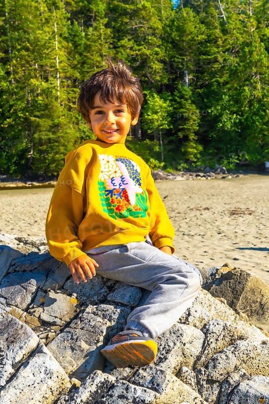 Happy child enjoying summer vacation, sitting on a rock at the beach in tofino, vancouver island, british columbia, canada, surrounded by lush green forest