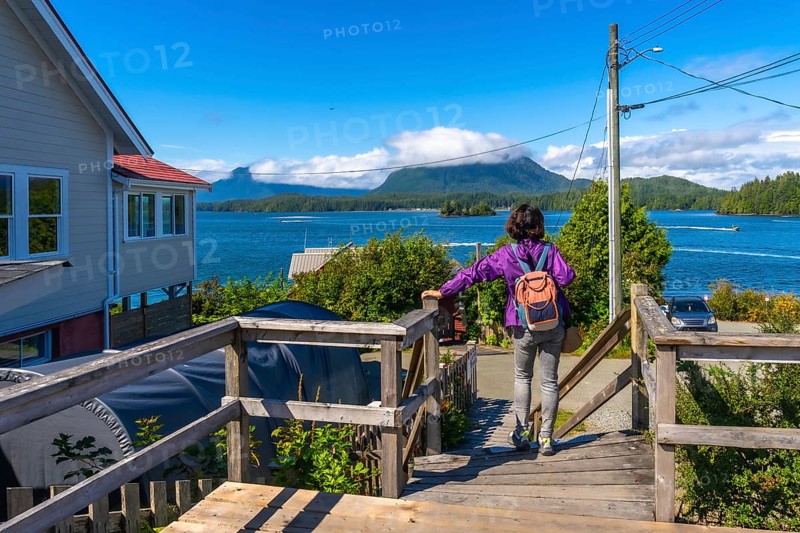 Female tourist with backpack walking down wooden stairs enjoying scenic view of tofino inlet, mountains, boats, and houses in tofino, vancouver island, british columbia, canada, during summer
